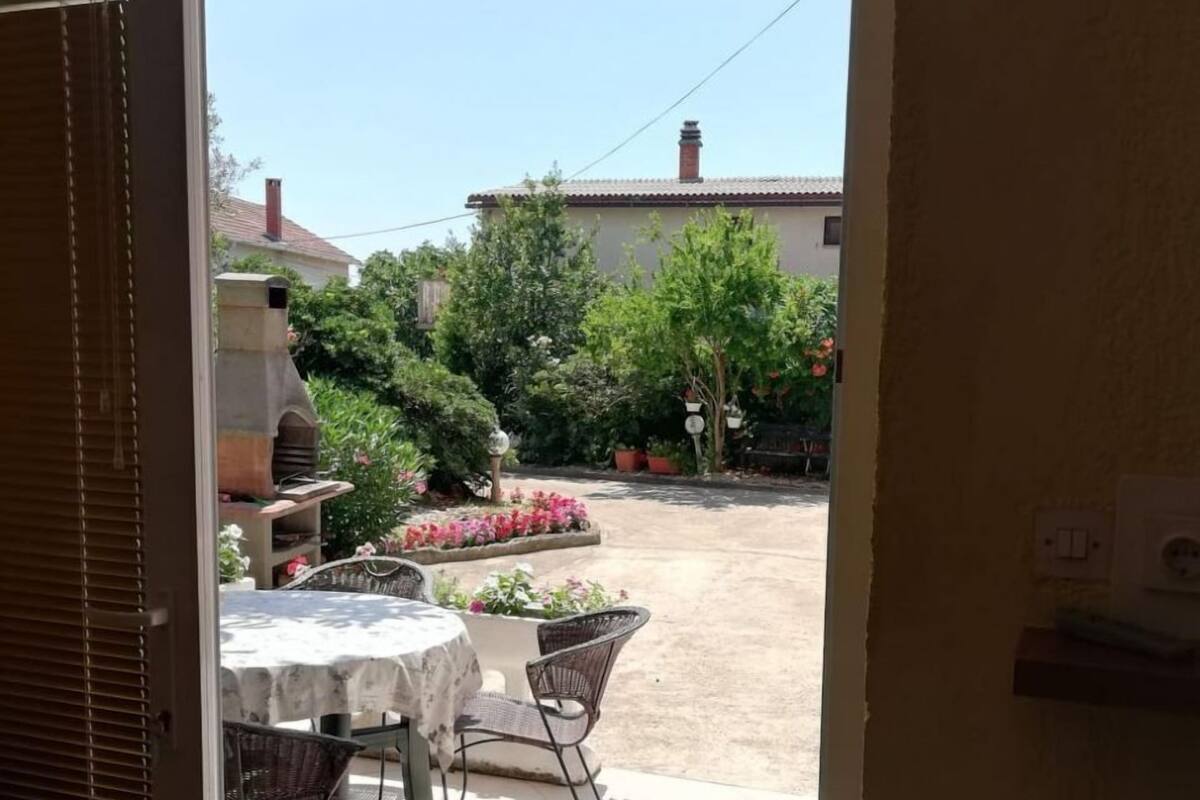 A patio with a table and chairs, a door leading outside, and a view of a garden with various plants and a building in the background.