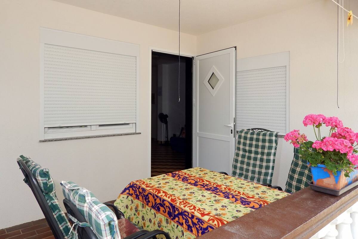 A room with a bed, a chair, a table with a colorful tablecloth, and a potted plant with pink flowers.