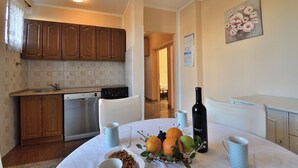 A kitchen with wooden cabinets, a dishwasher, a dining table with fruit and a bottle of wine, and a painting on the wall. - Apartment Nadica (Fazana)
