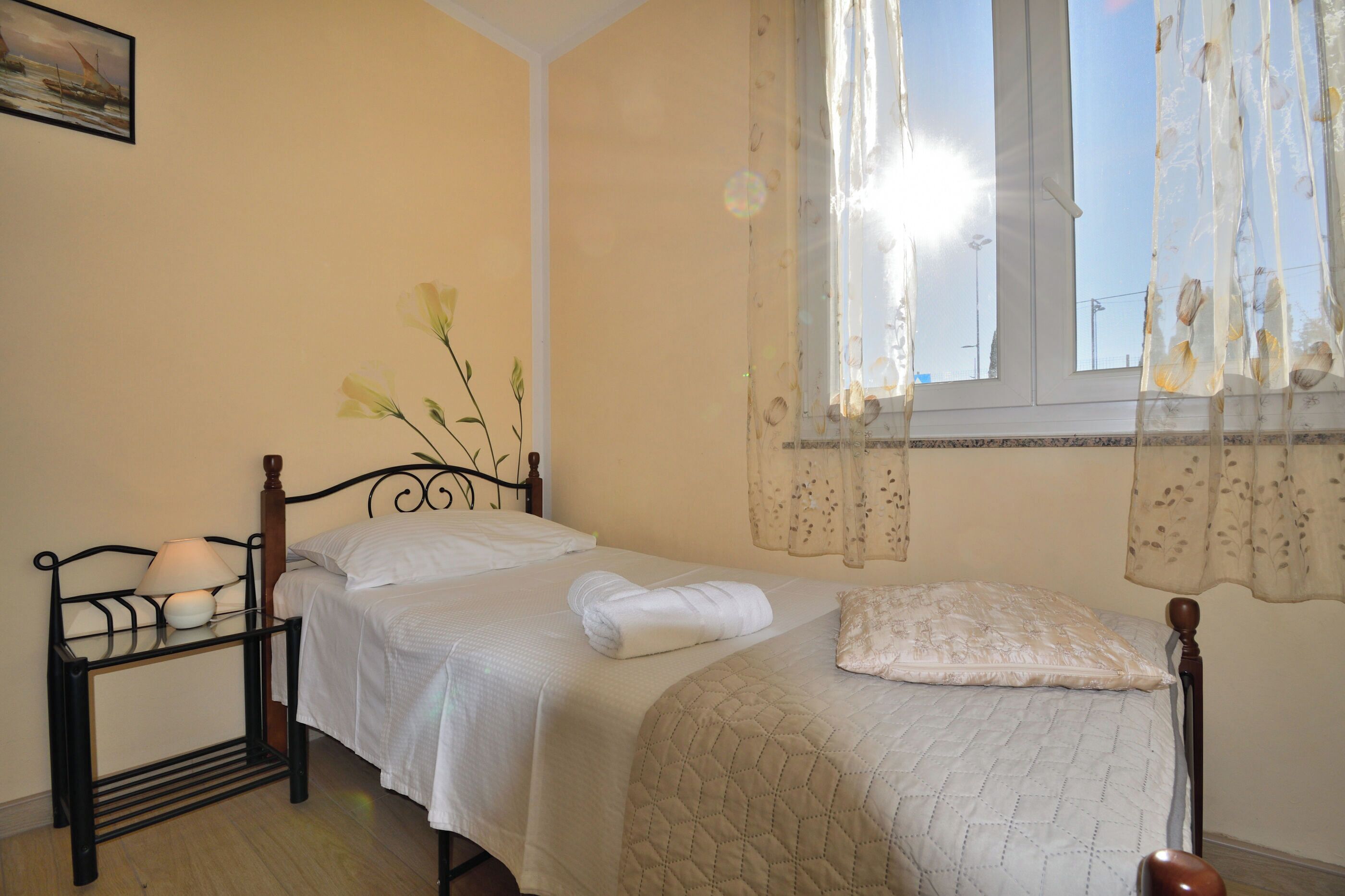 A neatly made bed with a quilted cover, a bedside table with a lamp, and a window with sheer curtains. - Apartment Nadica (Fazana)