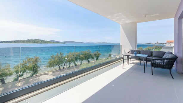 Ein Balkon mit Glasgeländer, einem Tisch und einer Couch mit Blick auf ein Gewässer und viel Grün.