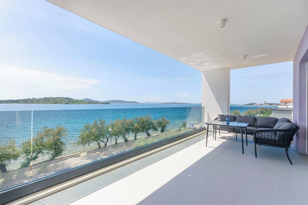 Un balcon doté d’une balustrade en verre, d’une table et d’un canapé, offrant une vue sur une étendue d’eau et la verdure.