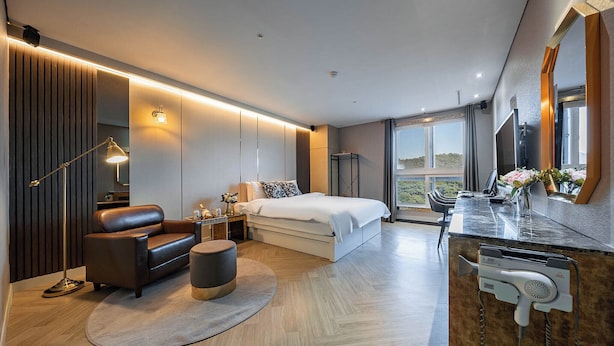 A modern hotel room with a large bed, a sitting area with a sofa and armchair, a small table with a lamp, and a hairdryer on the counter. - Dongtan Hotel Bella (Hwaseong)