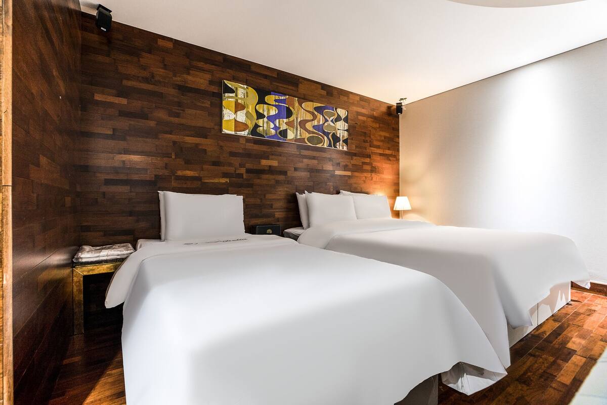 A hotel room with two beds, wooden paneling on the walls, a modern abstract painting, and a nightstand with a lamp.