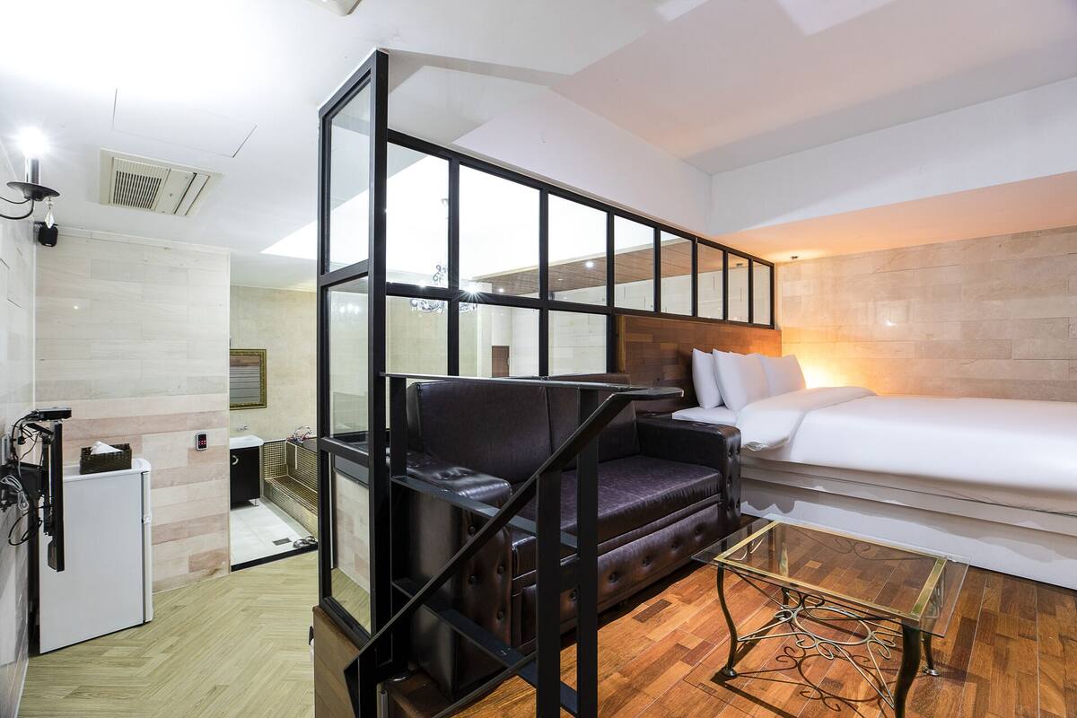 A modern hotel room with a glass partition, a bed, a seating area, and a small table.