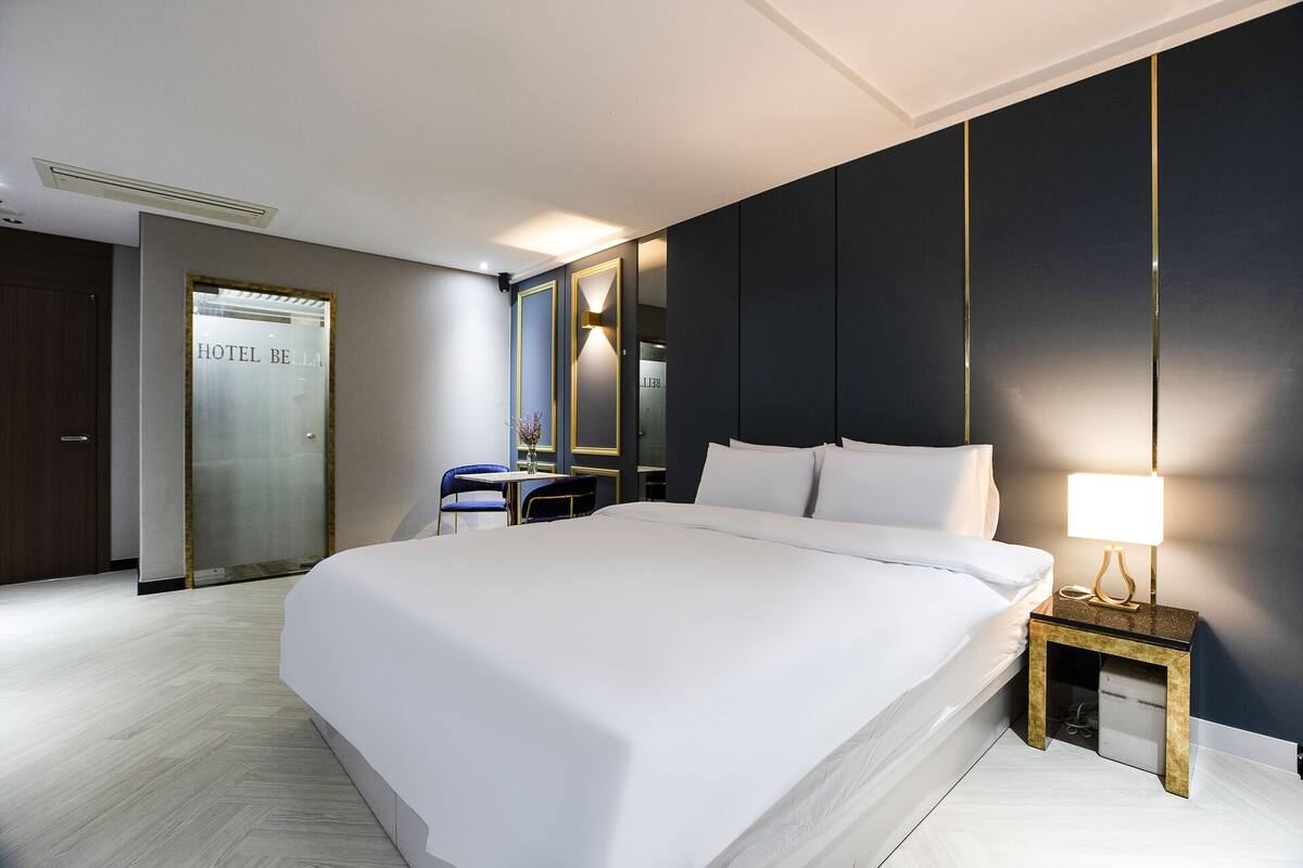 A modern hotel room with a large bed, a bedside table with a lamp, a mirror, and a small table with a vase of flowers.