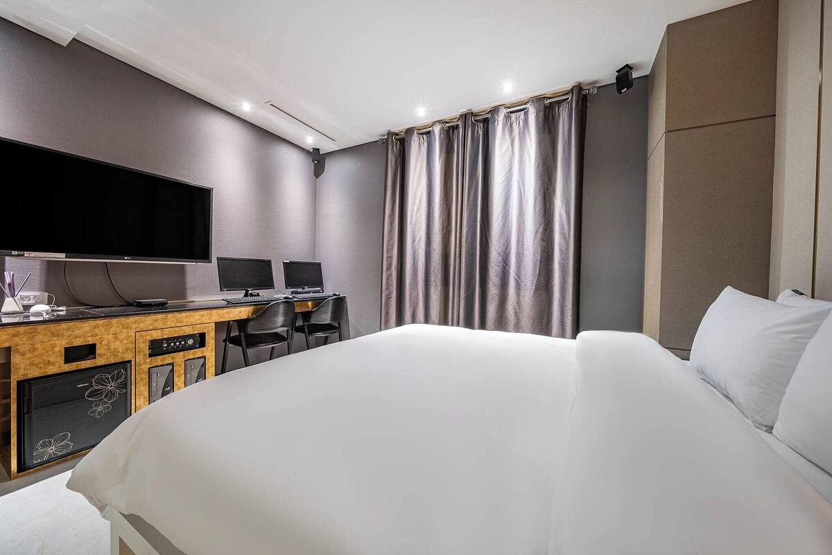 A hotel room with a large bed, a desk with two monitors, a flat-screen TV, and a minibar.