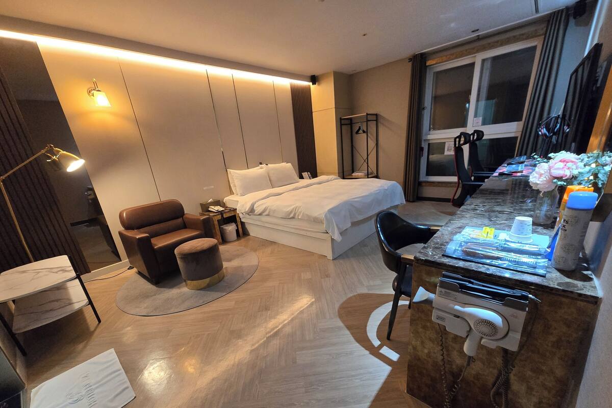A modern hotel room with a bed, a desk with a hairdryer, a chair, and a small table.