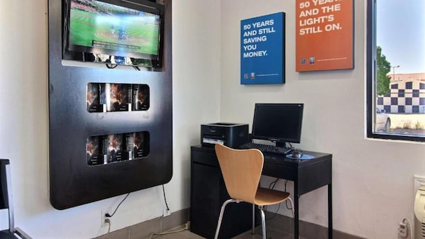 Una habitación con una televisión montada mostrando un evento deportivo, un escritorio con una computadora, una silla y dos carteles motivacionales en la pared.