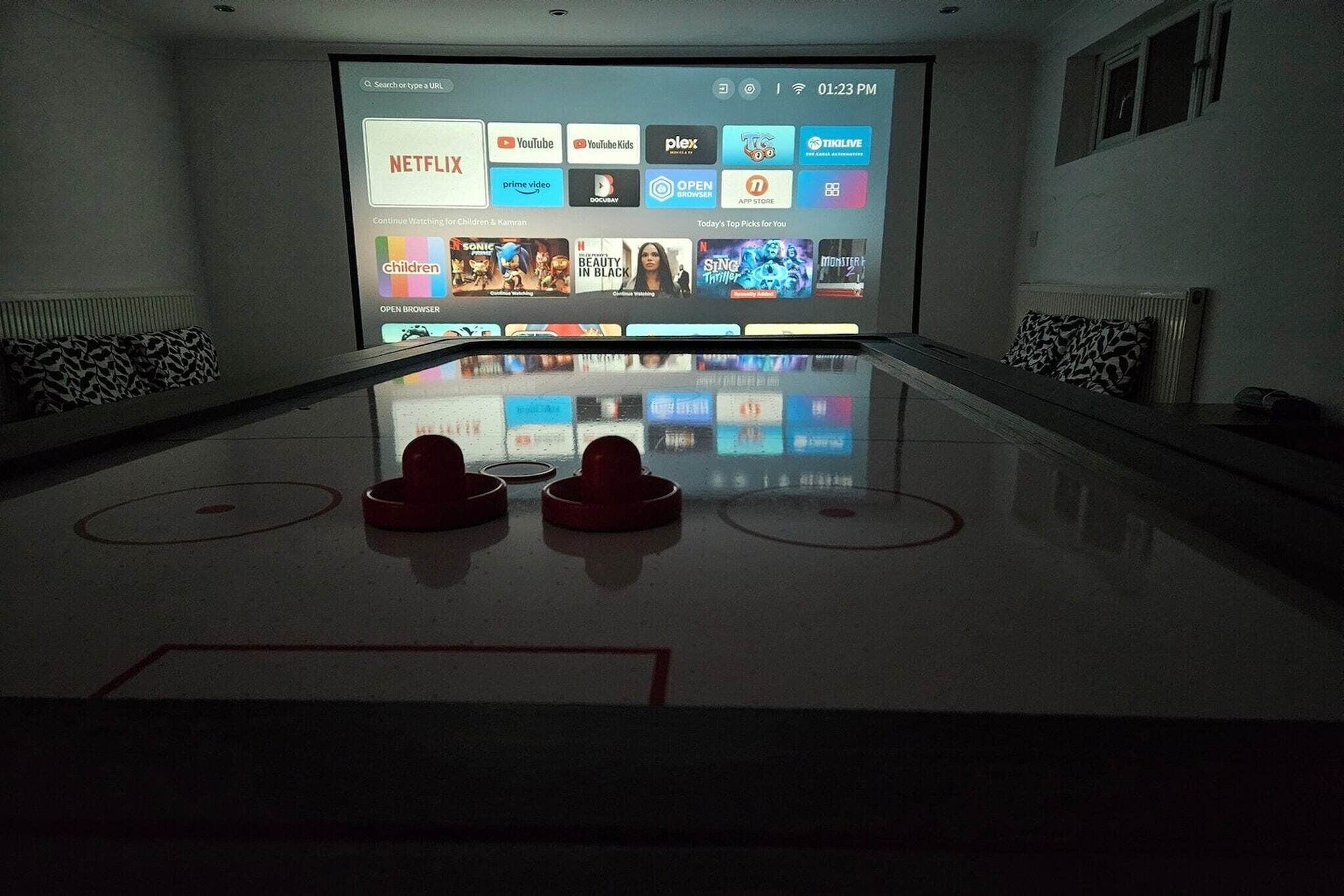 Ett rum med en stor, platt-TV som är monterad på väggen och som visar olika streamingtjänster. På golvet ligger två röda, runda föremål, eventuellt en del av ett spel eller en aktivitet. Rummet har en minimalistisk design med en vit vägg och en bänk med mönstrade kuddar.