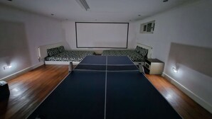 Ein Zimmer mit einer Tischtennisplatte, einer Couch mit gemusterten Kissen und einer Projektionsleinwand an der Wand.