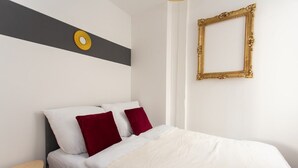 Una cama bien hecha con almohadas blancas y rojas, una pared gris y blanca con un adorno circular dorado, y un espejo con marco dorado en la pared.