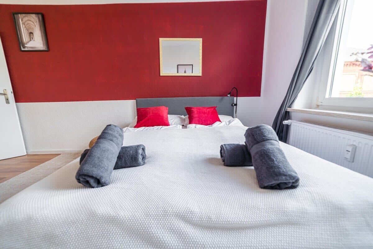 Een net opgemaakt bed met opgerolde handdoeken, een rode muur en een raam met gordijnen.
