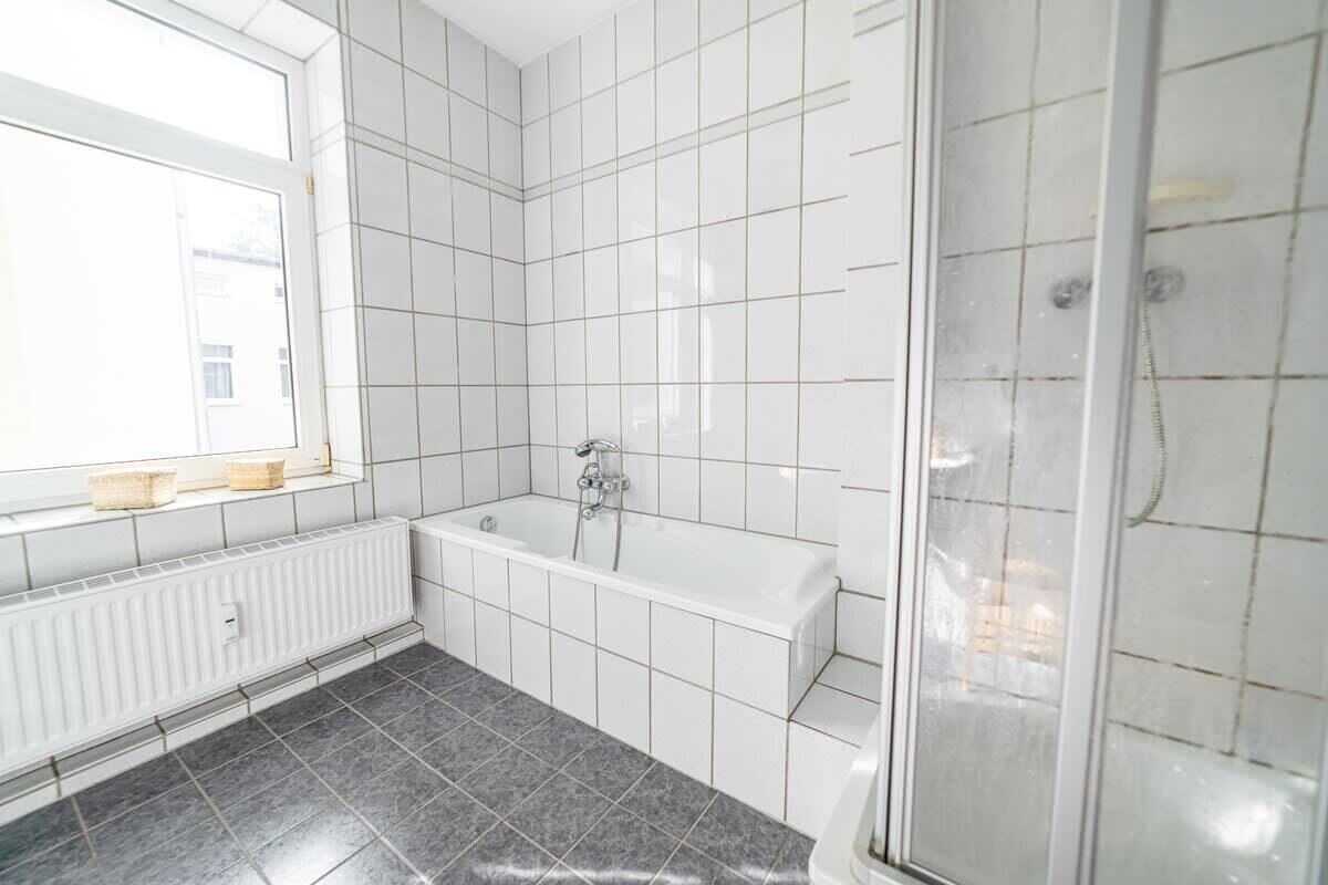 A bathroom with white tiles, a bathtub, a shower, and a window with baskets on the sill.