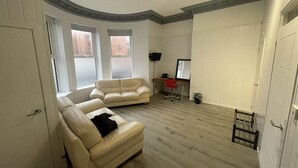 A room with a beige sofa, a desk with a computer, and a window with a view of buildings. - Standard Double Room (Belfast)