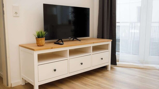 A white TV stand with a flat-screen TV, a small plant, and a remote control. - FullHouse Apartments AB5 Medium (Halle (Saale))