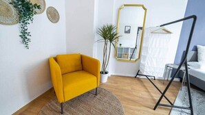 A modern living room with a yellow armchair, a round rug, a potted plant, a large mirror, and a black swing. - FullHouse | Erfurt | M18-3 | Caribbean Dream (Erfurt)