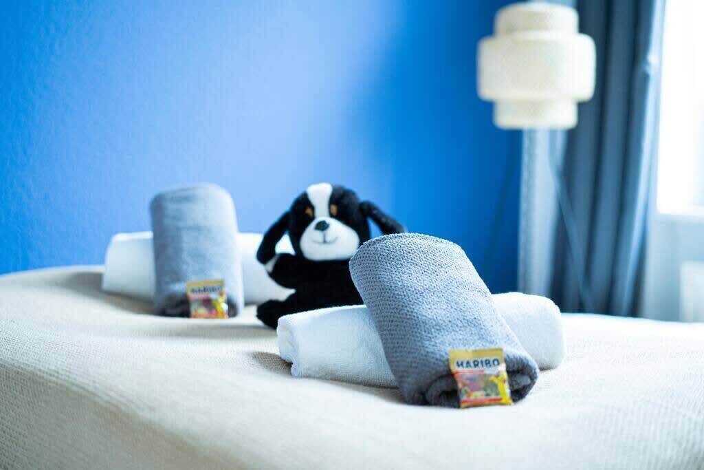 A plush toy dog on a bed with rolled towels and a bottle of hand sanitizer.