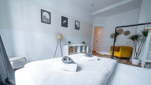 A modern hotel room with a large bed, a yellow armchair, and a white console table with plants. - FullHouse | Erfurt | M18-3 | Caribbean Dream (Erfurt)