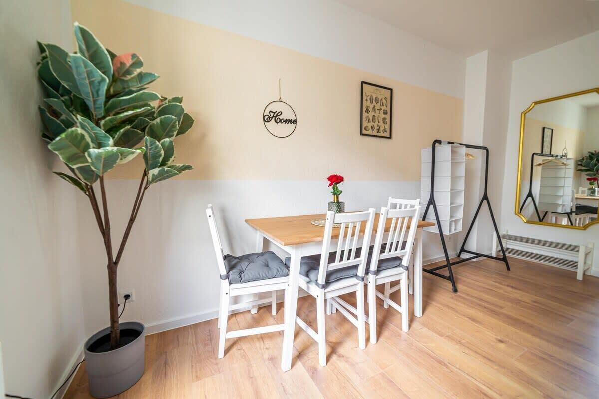 Een eetgedeelte met een houten tafel en witte stoelen, een potplant, een lijst met een foto en een spiegel met gouden lijst.