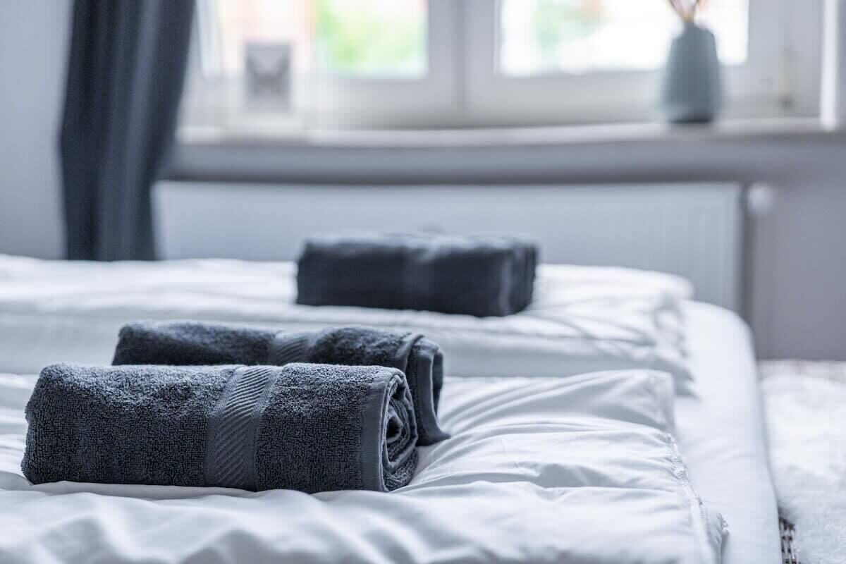 A neatly made bed with white bedding and two rolled towels placed on it.