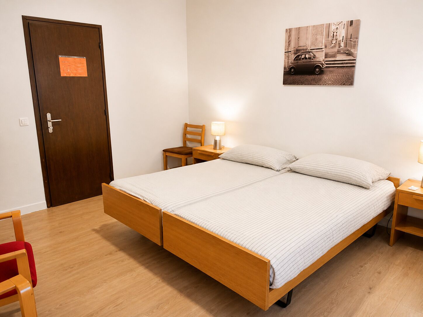 A single-bed room with a wooden frame, a small bedside table, a chair, a wall-mounted lamp, and a door with a notice.
