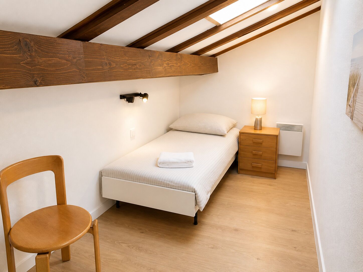 Een slaapkamer met een schuine kap, een bed, een nachtkastje, een stoel en een wandlamp.