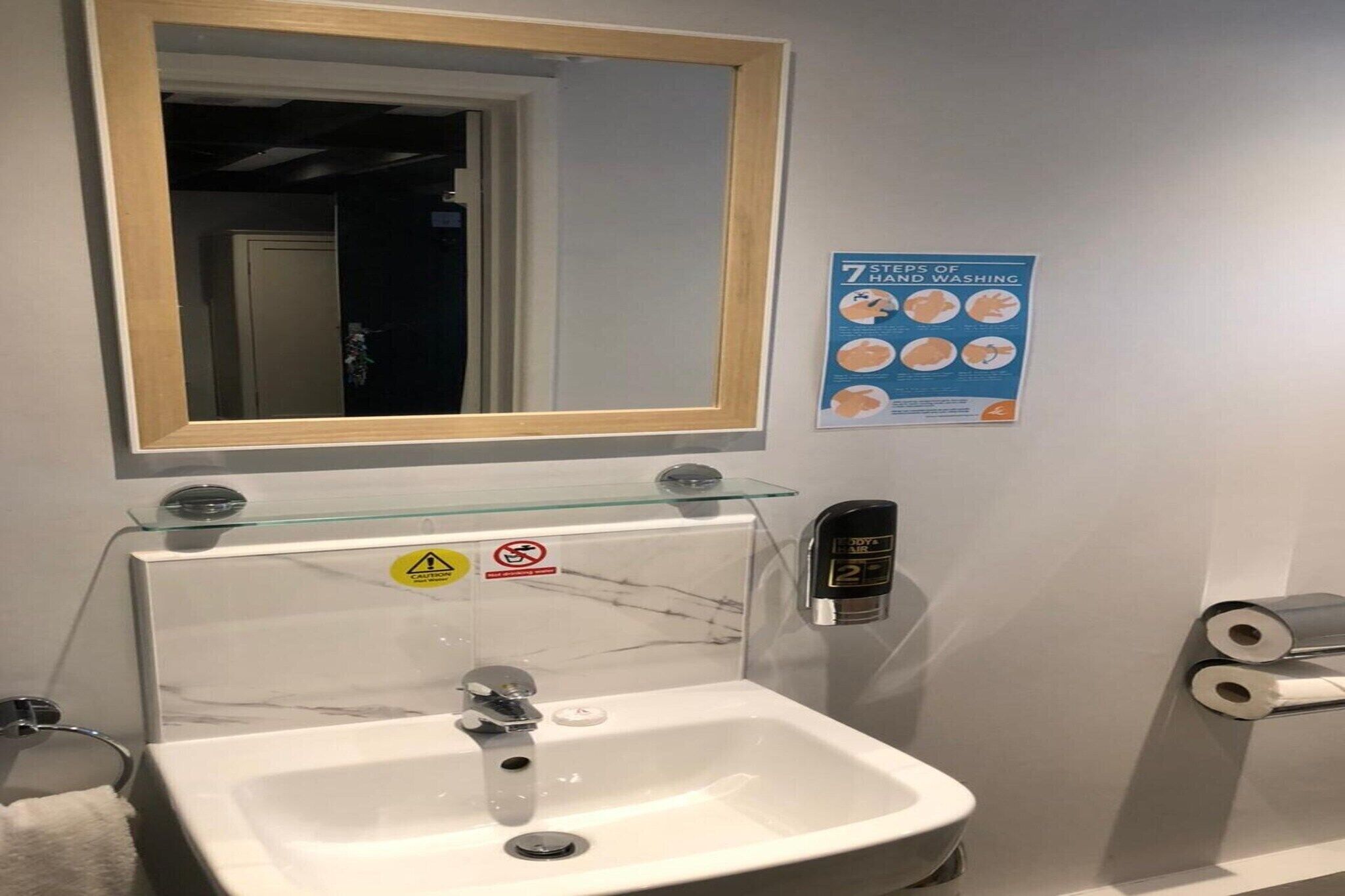 A bathroom with a sink, a mirror, a hand sanitizer dispenser, and a poster on the wall.