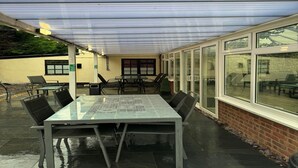 Un patio techado al aire libre con una mesa y sillas de vidrio, una pared de ladrillo y ventanas amplias.