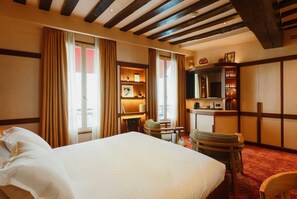 Quarto de hotel com cama grande, vigas de madeira no teto, escrivaninha com luminária e estante de livros.