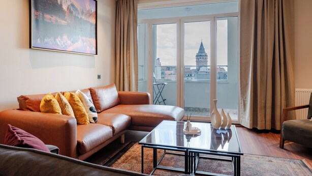 A modern living room with a leather sofa, a glass coffee table, and a large window offering a city view. - The Best in Beyoglu–Galata Tower View – Elevator – Renovated Interior – Balcony (İstanbul)