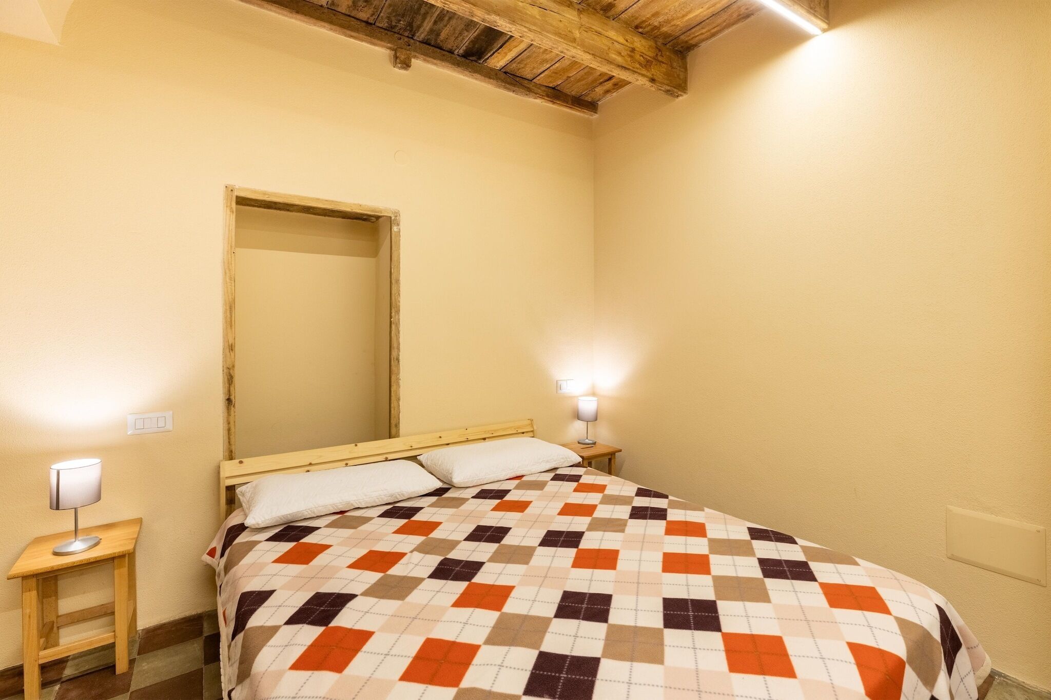 Un lit simple avec une couvre-lit à carreaux, une tête de lit en bois, une table de chevet avec une lampe et un miroir accroché au mur.