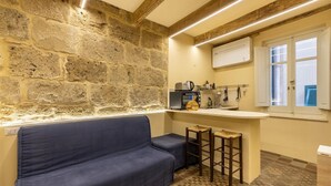 Une pièce chaleureuse avec un mur en pierre, un canapé, un petit coin bar avec des tabourets, un four à micro-ondes et une fenêtre avec des stores.