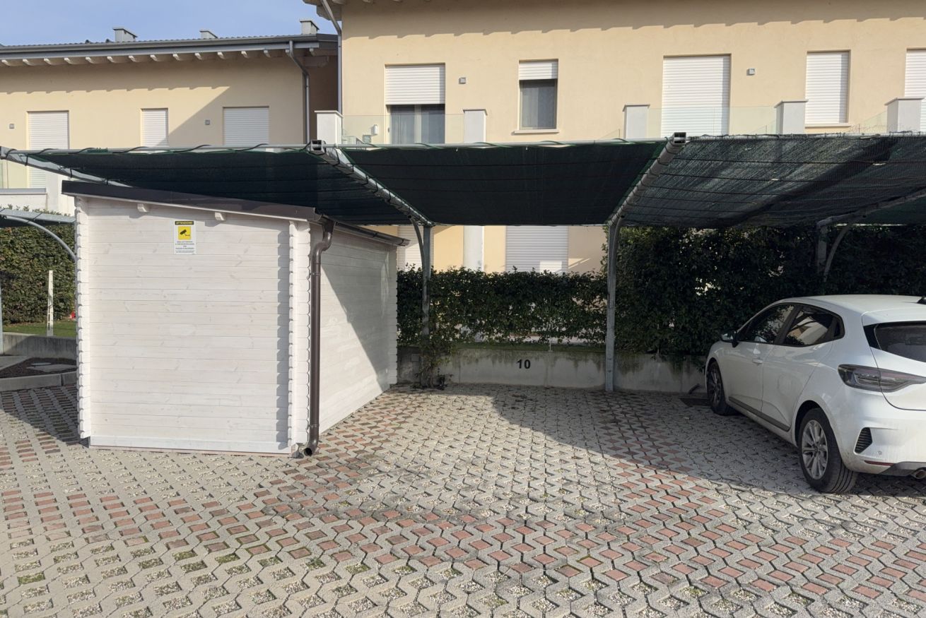 Un espace de stationnement couvert avec une voiture blanche, un petit abri de jardin et un bâtiment avec des fenêtres en arrière-plan.