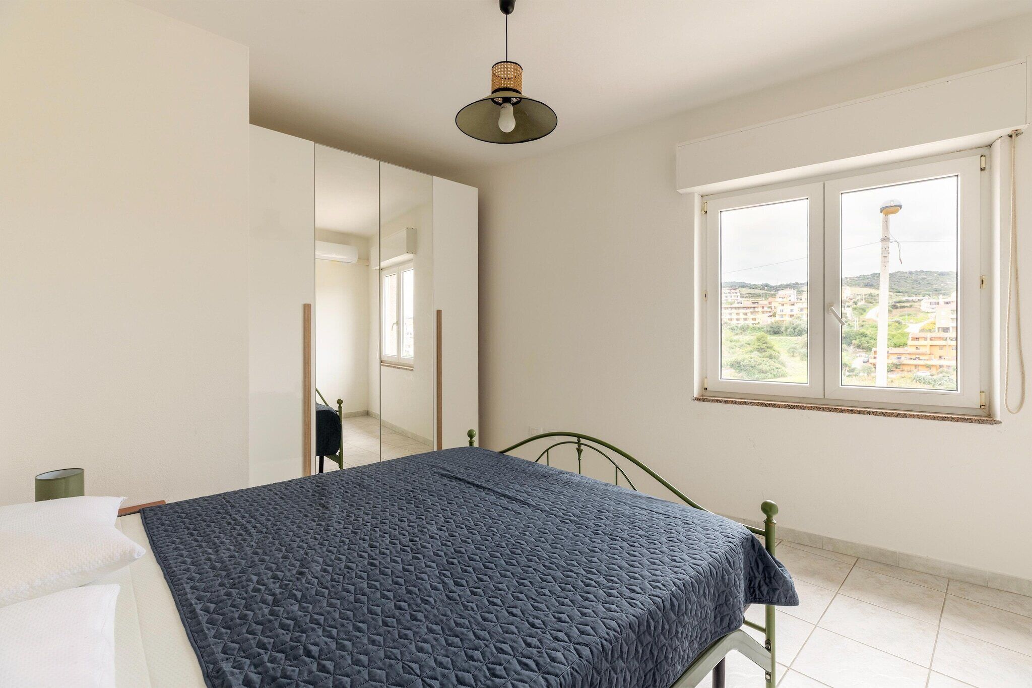 A bedroom with a bed, a window with a view, a mirror, and a hanging light fixture.