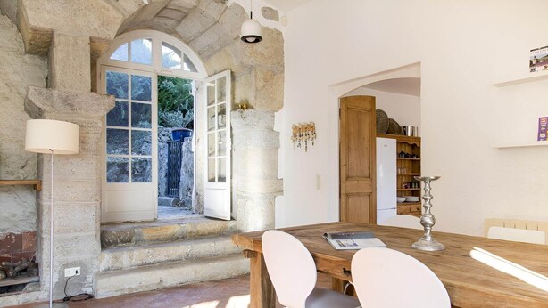 Un intérieur rustique avec des murs en pierre, une table à manger en bois, des chaises blanches et une cheminée en pierre.