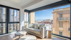 Ein modernes Wohnzimmer mit einer Couch und einem runden Tisch, großen Fenstern mit Blick auf die Stadt und einem Balkon mit Geländer.