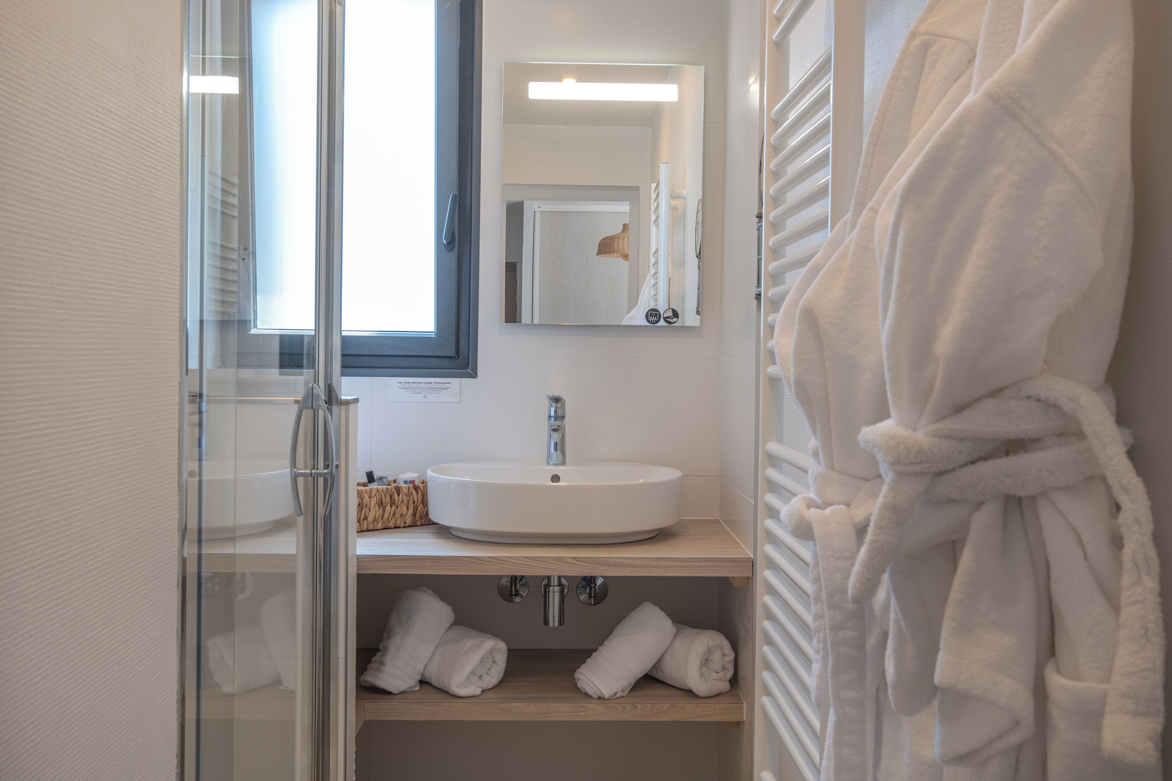 A bathroom with a glass shower enclosure, a white sink, and a mirror above it. There are towels on the shelves and a robe hanging on the wall.