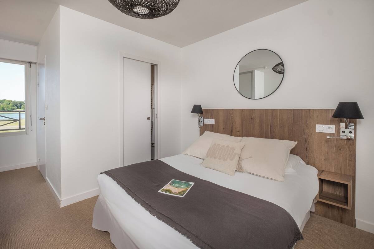 Une chambre à coucher avec un lit, une tête de lit en bois, un miroir rond, des lampes de chevet et une vue sur l’extérieur.