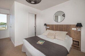 A bedroom with a bed, a wooden headboard, a round mirror, bedside lamps, and a view of the outdoors. - Hôtel de la Pointe du Cap Coz (Fouesnant)