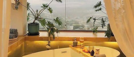 A modern bathroom with a freestanding bathtub, potted plants, and a city view through a large window.