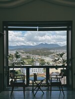 Uma varanda com duas cadeiras e uma mesa, com vista para uma paisagem urbana com montanhas ao longe.