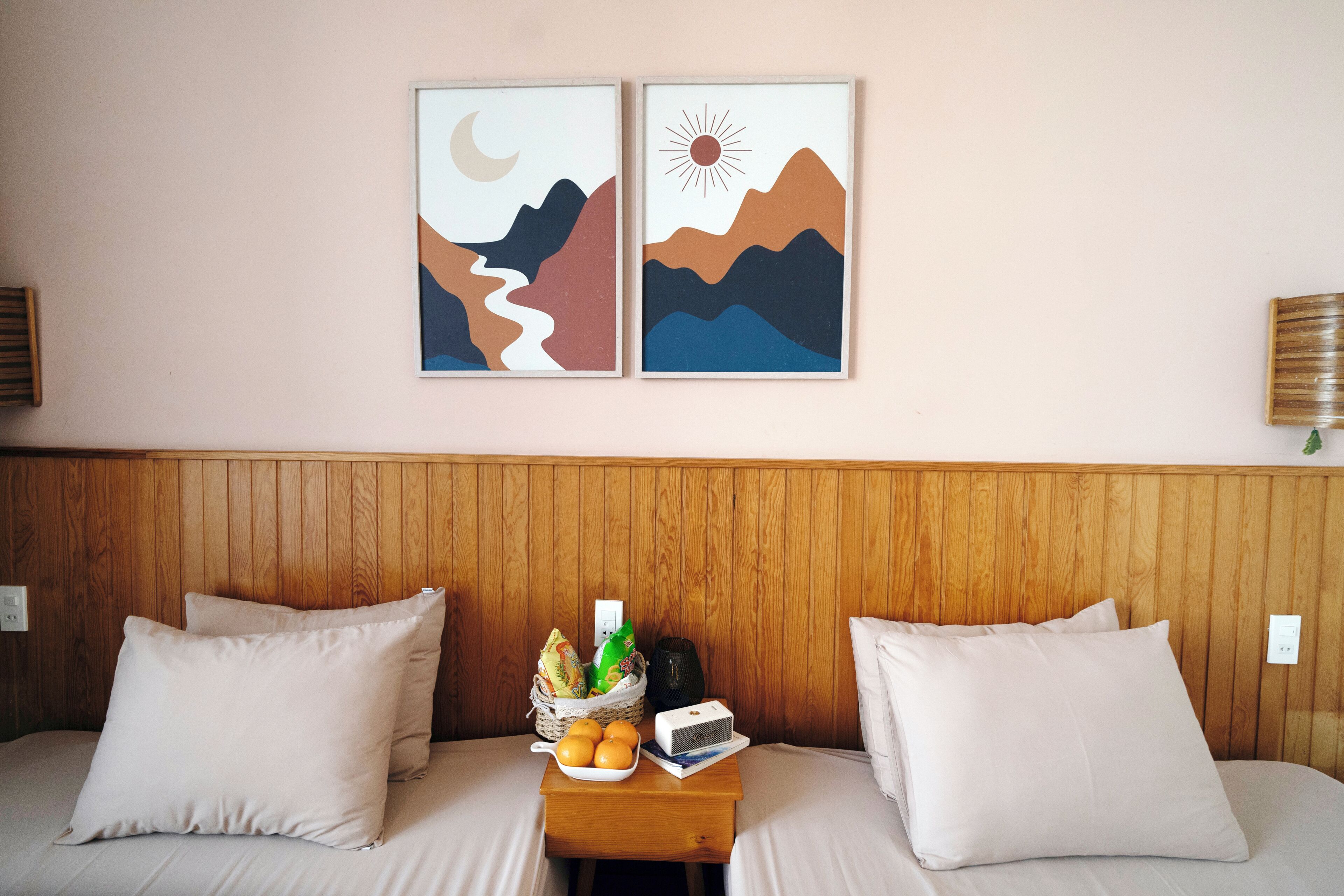 Habitación de hotel con cabecera de madera, dos almohadas blancas, una mesita de noche con una cesta de frutas y dos obras de arte enmarcadas en la pared.