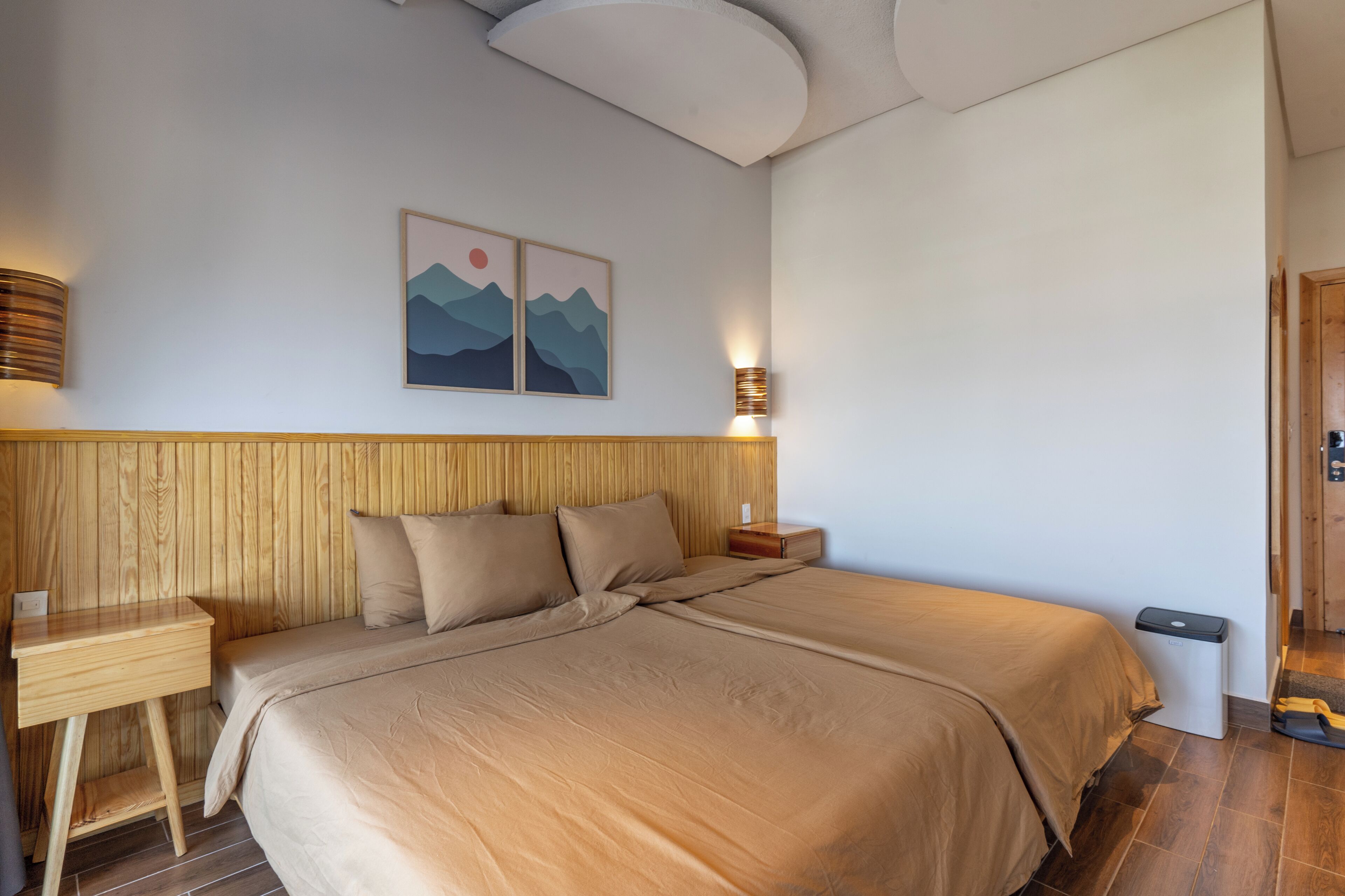 A bedroom with a wooden headboard, a bed with beige bedding, a bedside table, a wall-mounted lamp, and a framed picture above the bed.