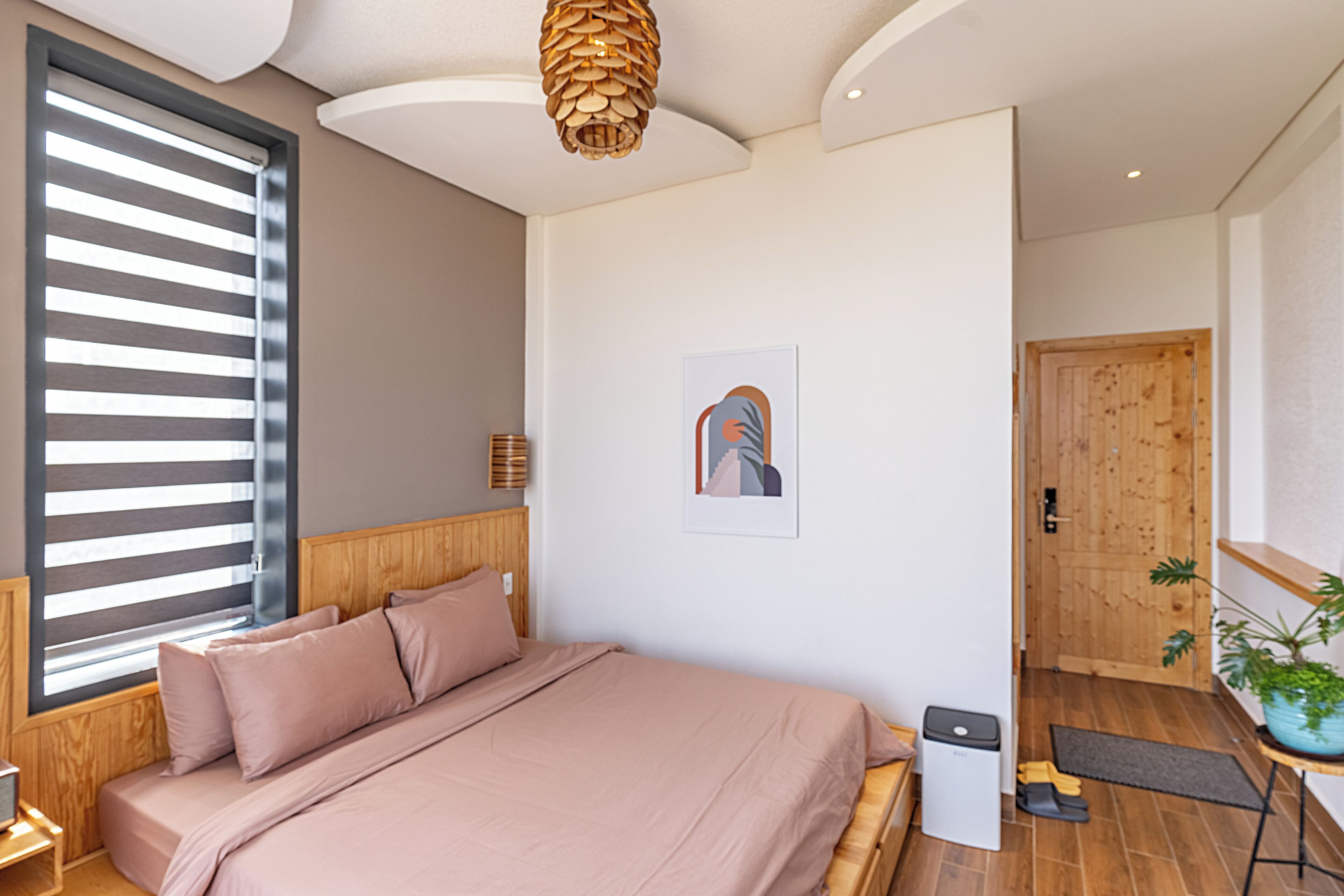 Un dormitorio moderno con una cama, cabecera de madera, una ventana con persianas, un cuadro colgado en la pared, una puerta de madera y una pequeña mesa con una planta.