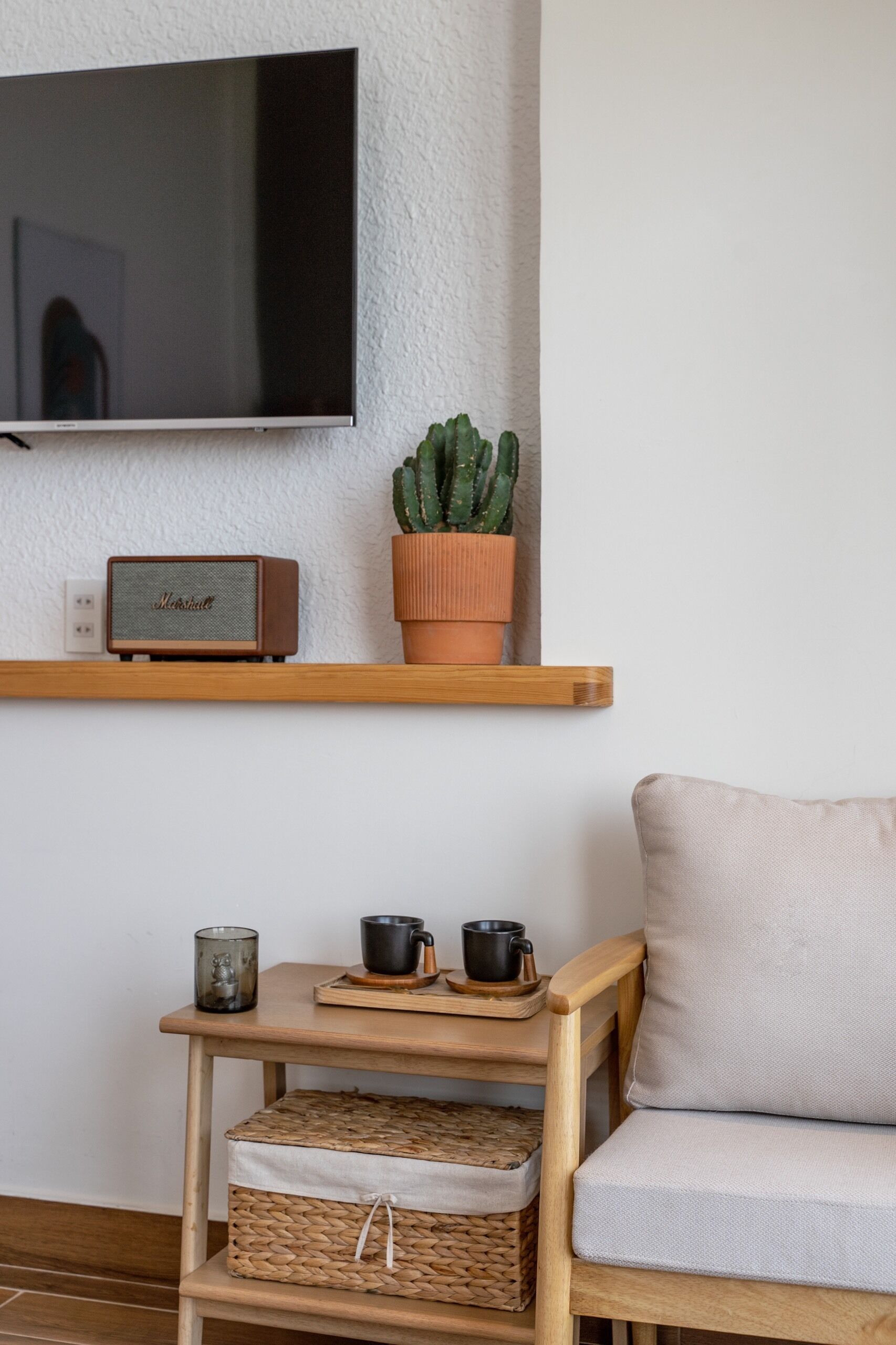 A living room with a flat-screen TV mounted on the wall, a wooden shelf with a cactus and a vintage radio, a small wooden table with two black mugs, and a cushioned chair.