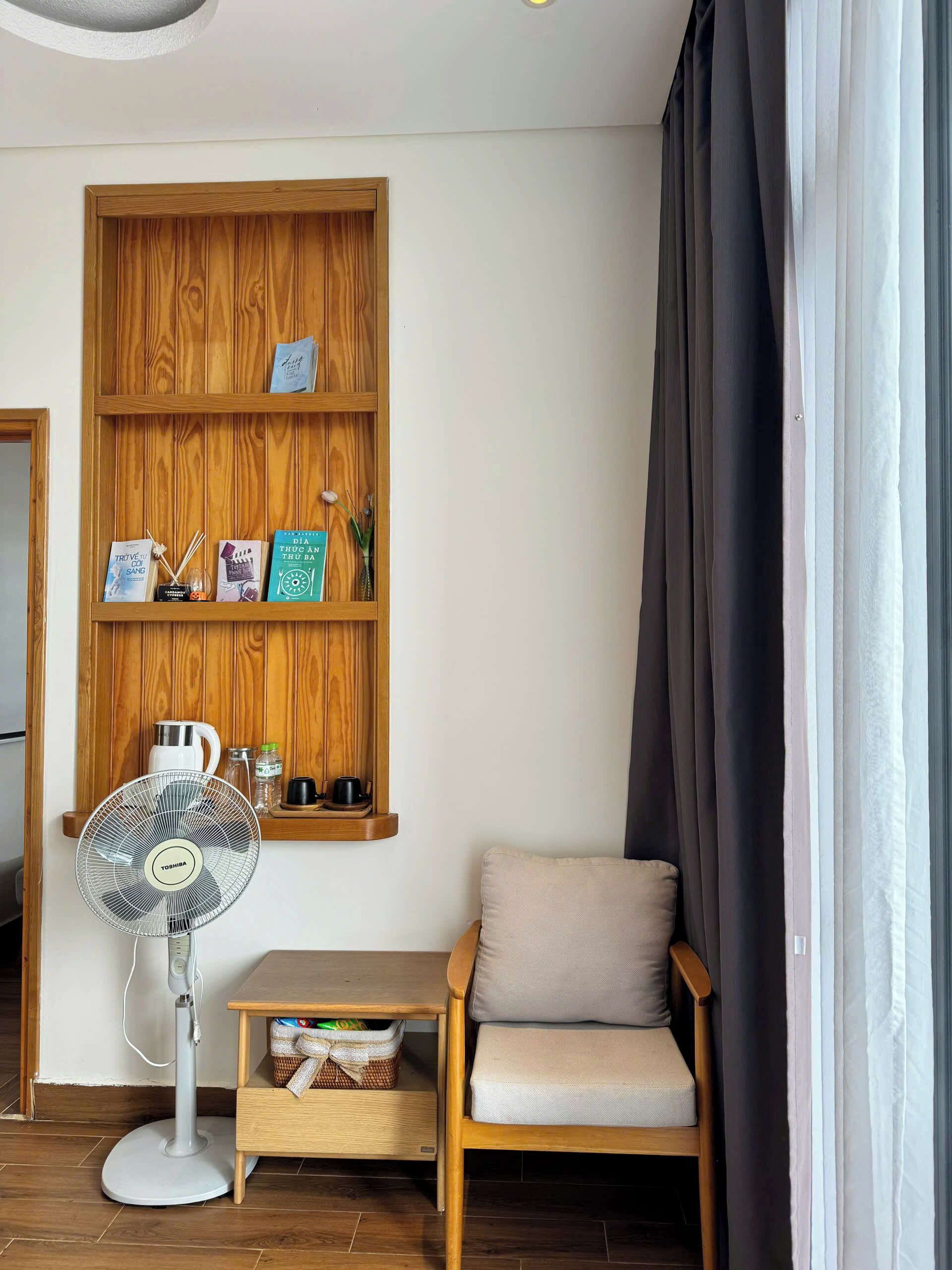A room with a wooden shelf unit, a fan, a chair, and a window with curtains.