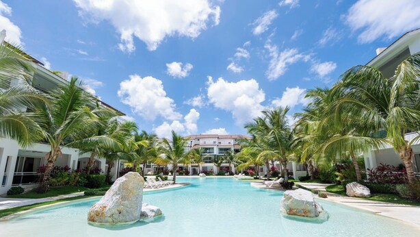 Zwembad van een resort met palmbomen, heldere lucht en een gebouw op de achtergrond.