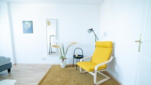 Ein helles Zimmer mit einem gelben Sessel, einem kleinen Tisch, einer Stehlampe und einem Spiegel an der Wand.