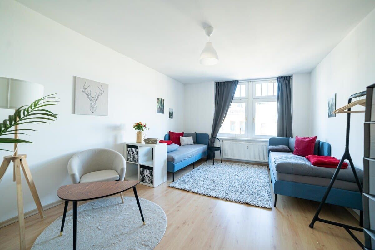 Una sala de estar moderna con un sofá, un sillón y una pequeña mesa. Hay una ventana con cortinas y una planta sobre un atril.